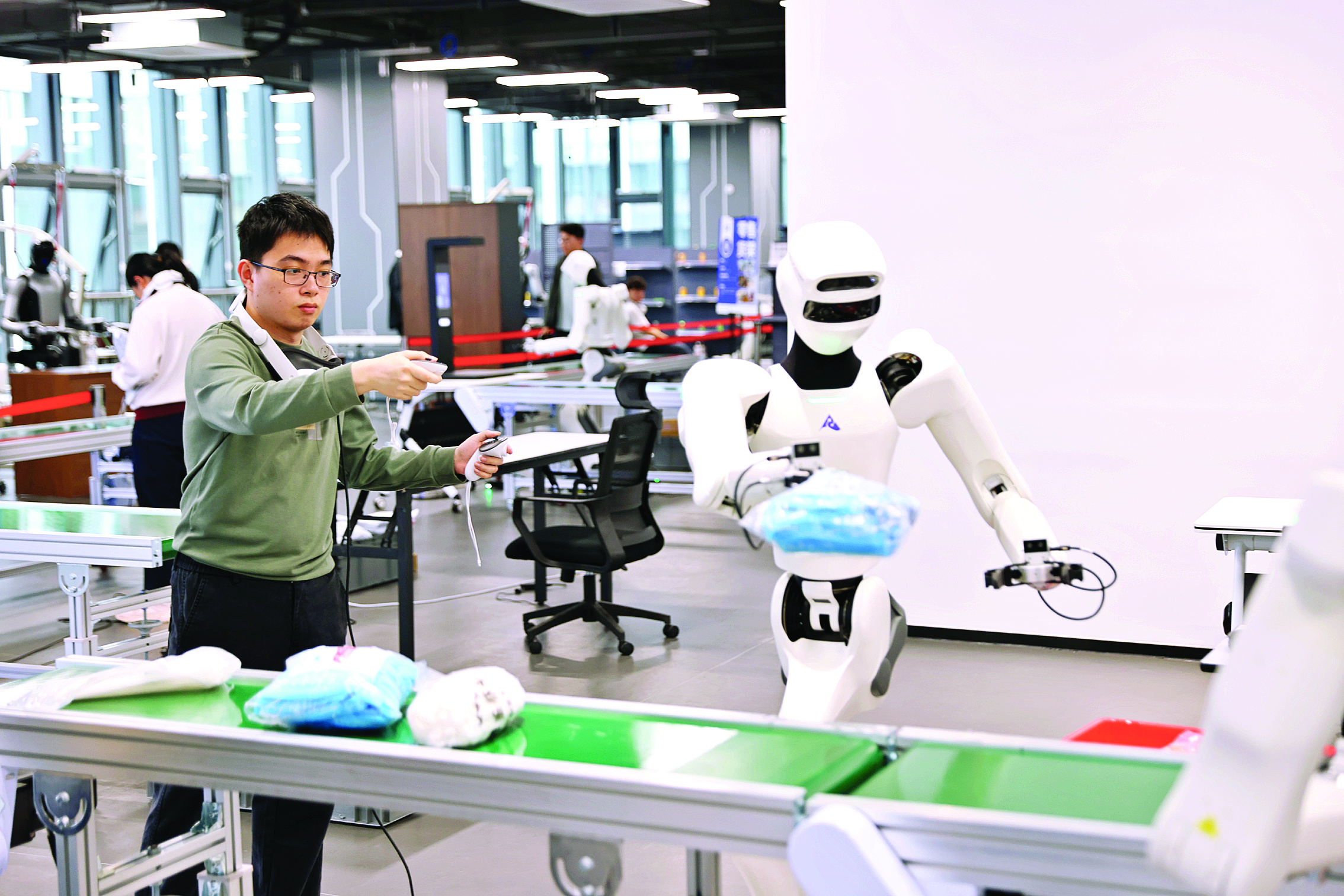     惠阳异构具身智能训练场里，机器人数据采集员在训练人形机器人模拟人形动作，在传送带上分拣物品。惠州日报记者李松权 汤渝杭 摄